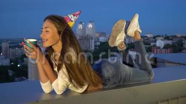 快乐的女孩戴着<strong>节</strong>日的帽子庆祝她的生日，吹<strong>进口</strong>哨，拿着纸杯，躺在屋顶上